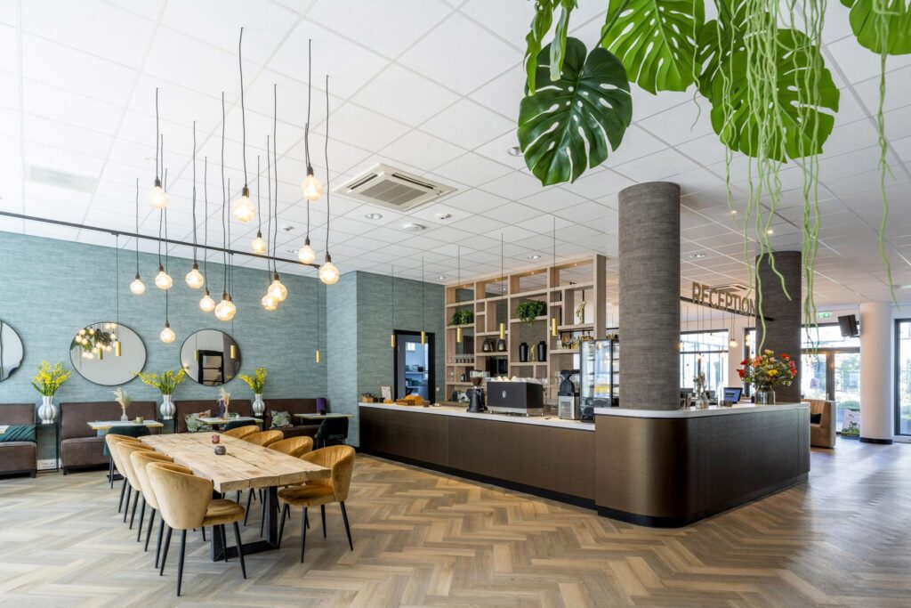 Modern hotel reception area with a wooden table, cozy seating, hanging lights, and decorative plants.